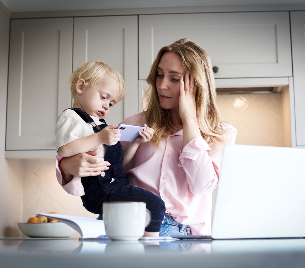 A parent holding a toddler while working at a laptop, hand to their temple, carrying the household's mental load